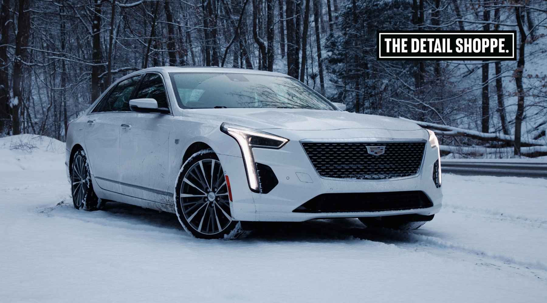A white Cadillac sedan with custom wheels parked on a snowy road, showing signs of road salt and grime on the lower panels. The backdrop features a winter forest, and a "The Detail Shoppe" logo appears in the top right corner.