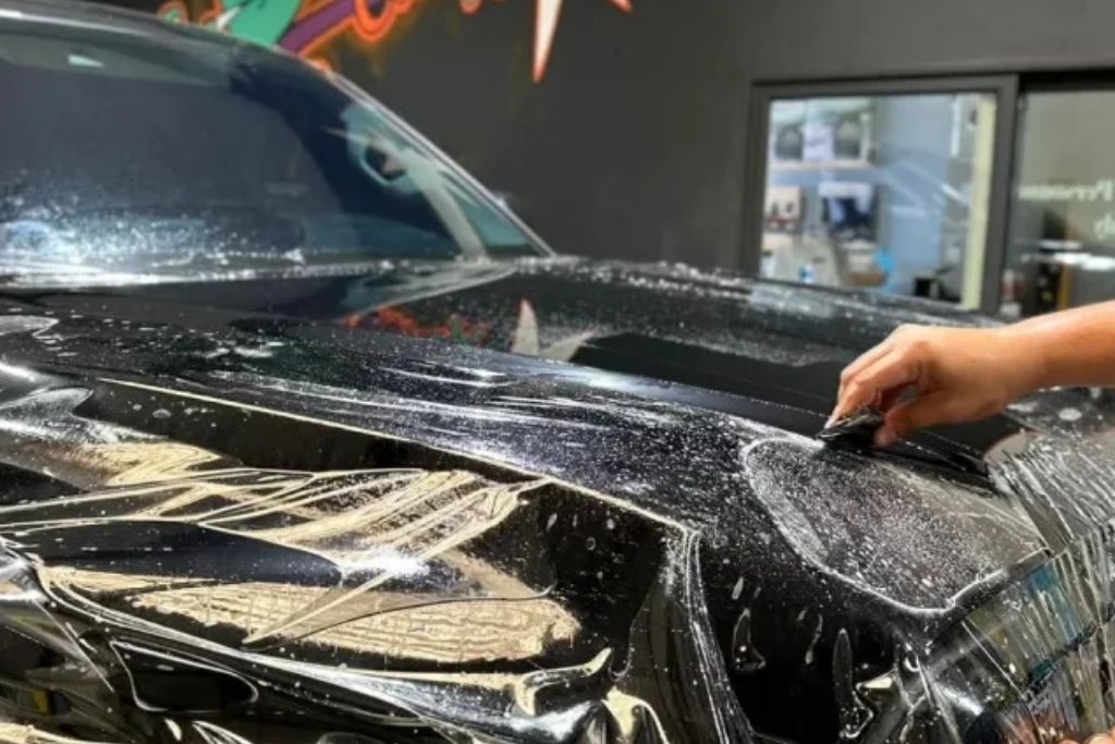 Technician smoothing out paint protection film on a black vehicle hood during installation.