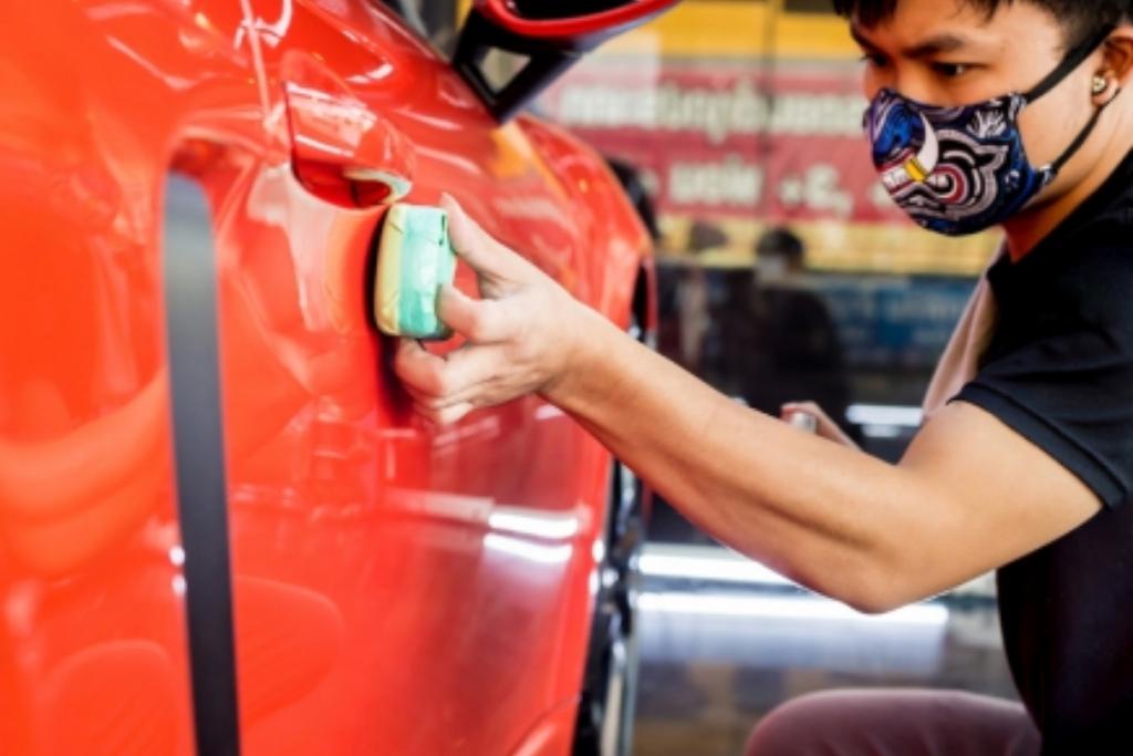 Auto detailing technician applying wax or protectant to a red car door with a foam applicator pad.