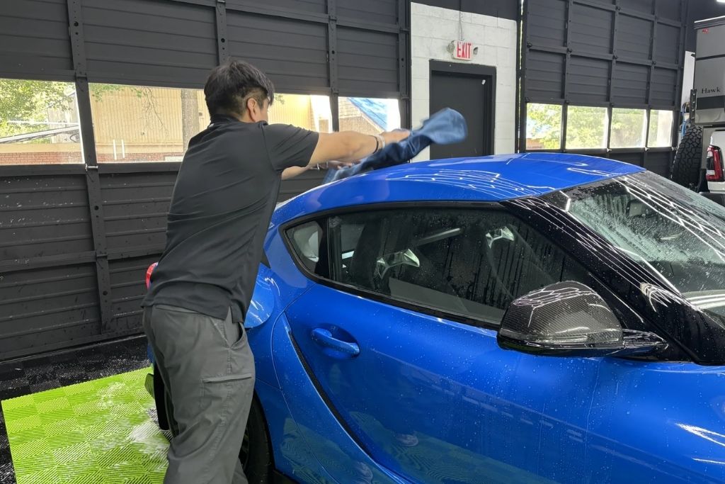 Technician drying a blue sports car after paint protection film installation at a detailing shop.