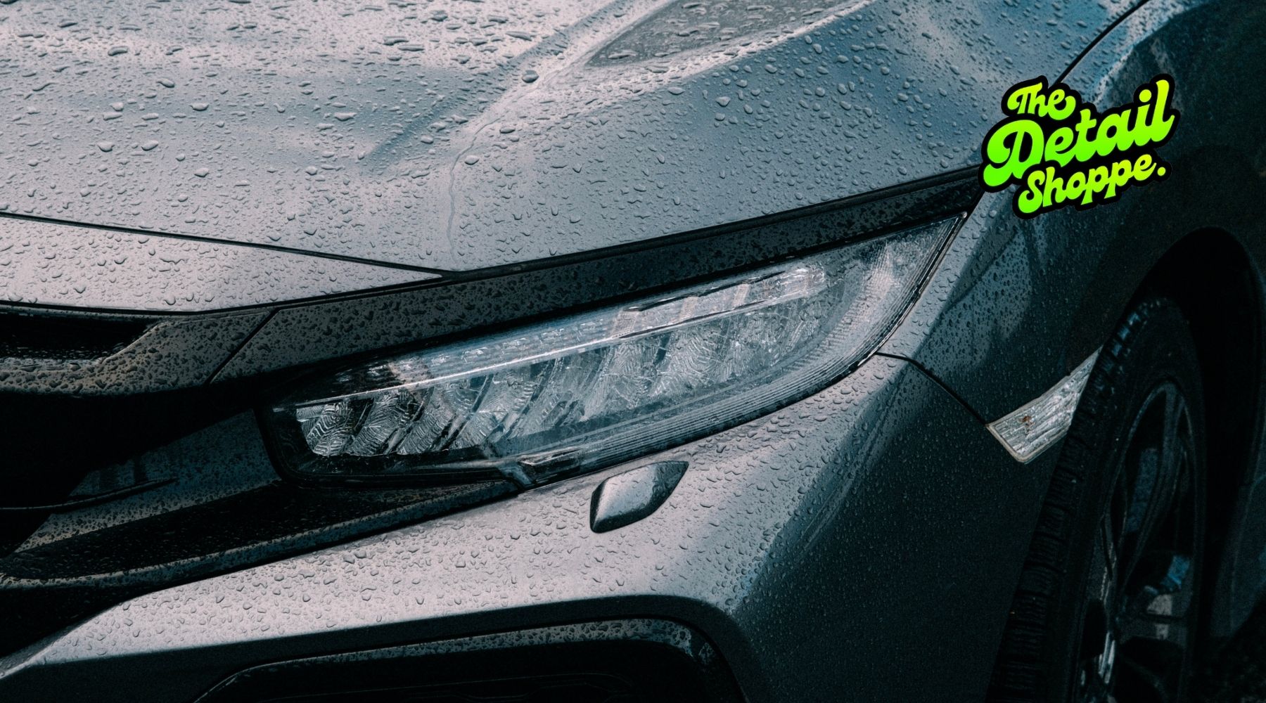 Close-up of a gray car covered in rain droplets highlighting the headlight and glossy paint surface.