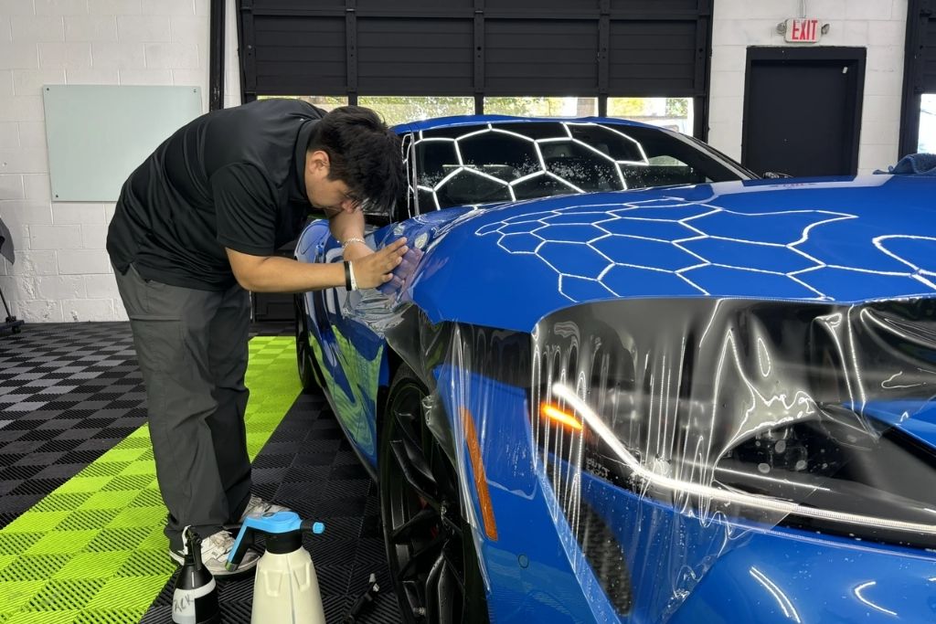 Technician installing paint protection film on a blue sports car inside a professional auto detailing shop.