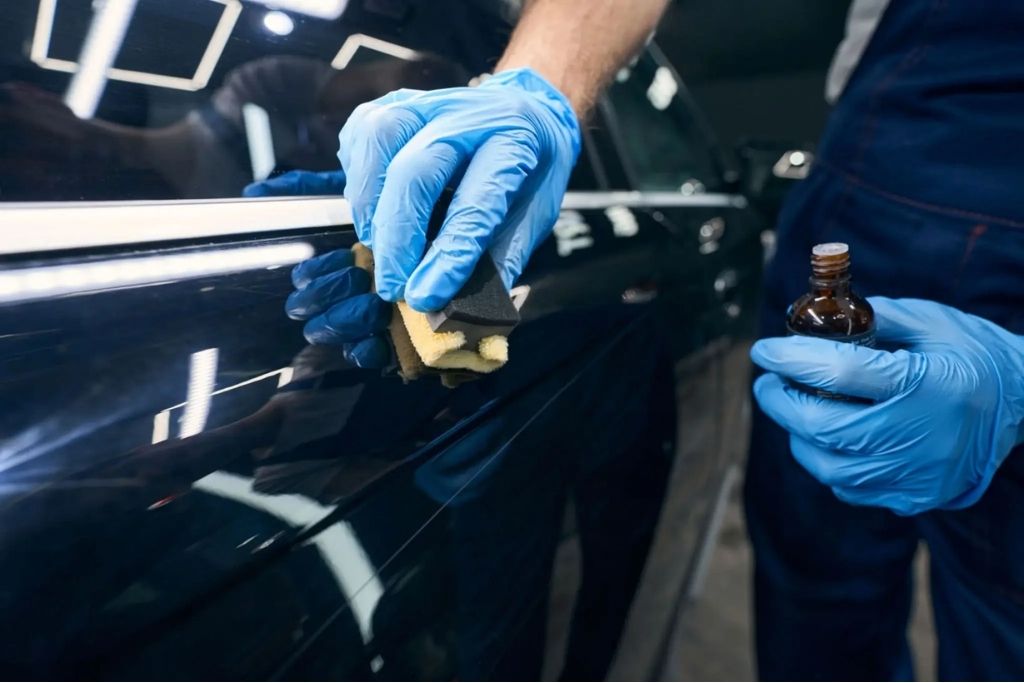 Technician wearing blue gloves applying ceramic coating to a black car door with an applicator pad.