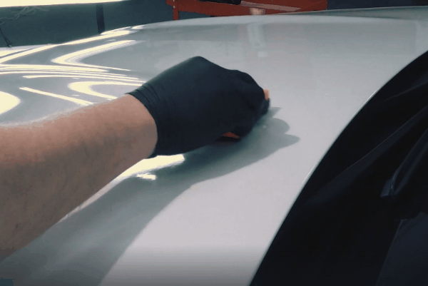 Technician applying ceramic coating to a white car surface using an applicator pad while wearing a black glove.